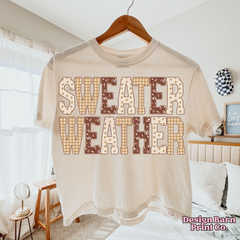 Puede incluir: Camiseta color crema con las palabras "SWEATER WEATHER" en una fuente decorativa. Las letras presentan patrones de cuadros y florales en tonos marrones y beige. La camiseta est&aacute; colgada en una percha de madera.