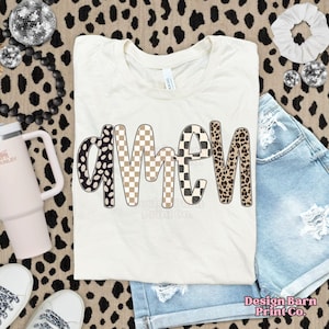 Op de afbeelding: Cr&egrave;mekleurig T-shirt met het woord "AMEN" in een speels, patroonachtig lettertype. De letters zijn voorzien van zwart-witte stippen, bruin-wit dambord, zwart-wit dambord en luipaardprint. Het shirt wordt gecombineerd met een spijkerbroekshort.