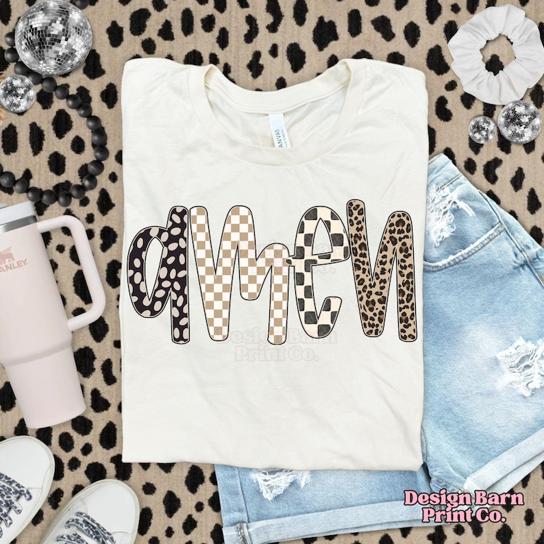 Op de afbeelding: Cr&egrave;mekleurig T-shirt met het woord "AMEN" in een speels, patroonachtig lettertype. De letters zijn voorzien van zwart-witte stippen, bruin-wit dambord, zwart-wit dambord en luipaardprint. Het shirt wordt gecombineerd met een spijkerbroekshort.