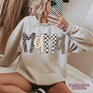 K&ouml;nnte beinhalten: Beiges Sweatshirt mit dem Wort "MAMA" in gro&szlig;en, gemusterten Buchstaben. Die Buchstaben zeigen schwarz-wei&szlig;e Punkte, ein beige-wei&szlig;es Schachbrettmuster und ein Leopardenmuster. Das Sweatshirt hat lange &Auml;rmel.