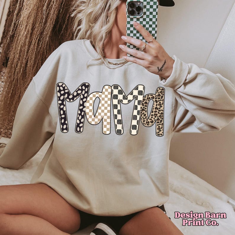 K&ouml;nnte beinhalten: Beiges Sweatshirt mit dem Wort "MAMA" in gro&szlig;en, gemusterten Buchstaben. Die Buchstaben zeigen schwarz-wei&szlig;e Punkte, ein beige-wei&szlig;es Schachbrettmuster und ein Leopardenmuster. Das Sweatshirt hat lange &Auml;rmel.