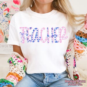 Pode incluir: Camiseta branca com a palavra "TEACHER" em estampa xadrez rosa e azul. A palavra "TEACHER" tem detalhes florais e la&ccedil;os. Faz parte de uma cole&ccedil;&atilde;o com tema de professores.