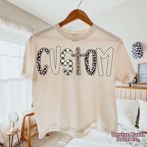 May include: Cream-colored t-shirt with the word "CUSTOM" in a bold, playful font. The letters feature a mix of patterns, including black and white polka dots, checkered, and leopard print. The shirt is hanging on a wooden hanger.