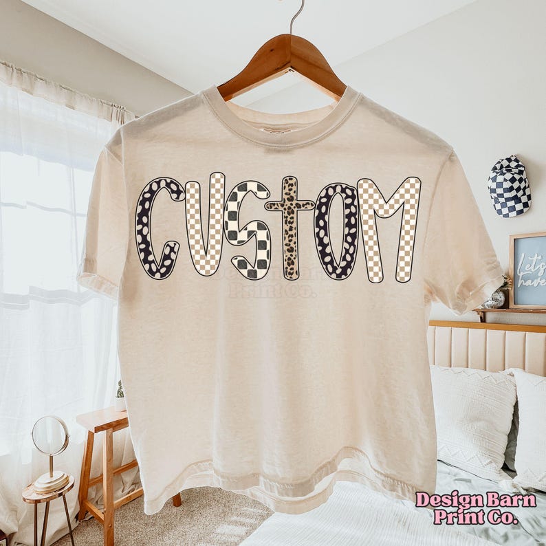 May include: Cream-colored t-shirt with the word "CUSTOM" in a bold, playful font. The letters feature a mix of patterns, including black and white polka dots, checkered, and leopard print. The shirt is hanging on a wooden hanger.