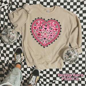K&ouml;nnte beinhalten: Ein beiges Sweatshirt mit einem gro&szlig;en Herzdesign, das mit rosa Blumen und schwarzen Herzformen gef&uuml;llt ist. Das Herz ist von einem dunkelrosa, gewellten Rand umgeben. Der Text "Design Barn Print Co." ist unten rechts zu sehen.