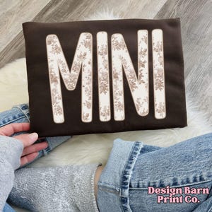 Puede incluir: Una sudadera marr&oacute;n oscuro con la palabra "MINI" en letras grandes con un estampado floral. La sudadera la sostiene una persona que lleva vaqueros azules y una sudadera gris. El texto "Design Barn Print Co." es visible en la esquina inferior derecha.
