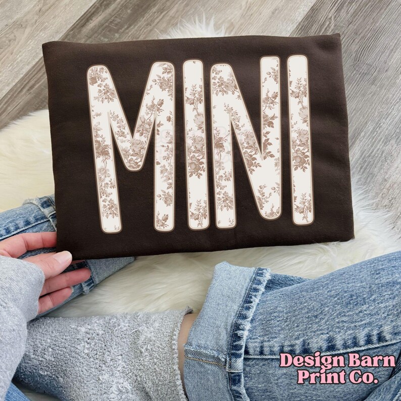 Puede incluir: Una sudadera marr&oacute;n oscuro con la palabra "MINI" en letras grandes con un estampado floral. La sudadera la sostiene una persona que lleva vaqueros azules y una sudadera gris. El texto "Design Barn Print Co." es visible en la esquina inferior derecha.