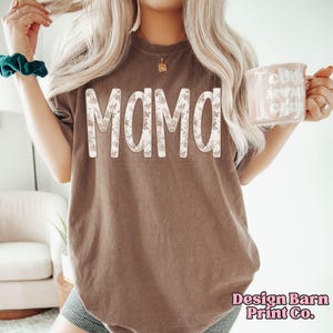 May include: Brown t-shirt with the word "MAMA" in large, floral-patterned letters. The shirt has a vintage look and is from Design Barn Print Co. The shirt is a casual style.
