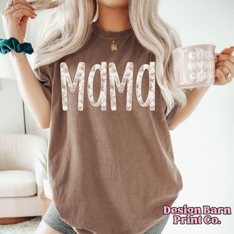 May include: Brown t-shirt with the word "MAMA" in large, floral-patterned letters. The shirt has a vintage look and is from Design Barn Print Co. The shirt is a casual style.