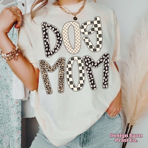 May include: Cream-colored t-shirt with the words "DOG MOM" in a bold, patterned design. The letters feature a mix of black and white polka dots, checkered patterns, and leopard print. The shirt is a casual style.