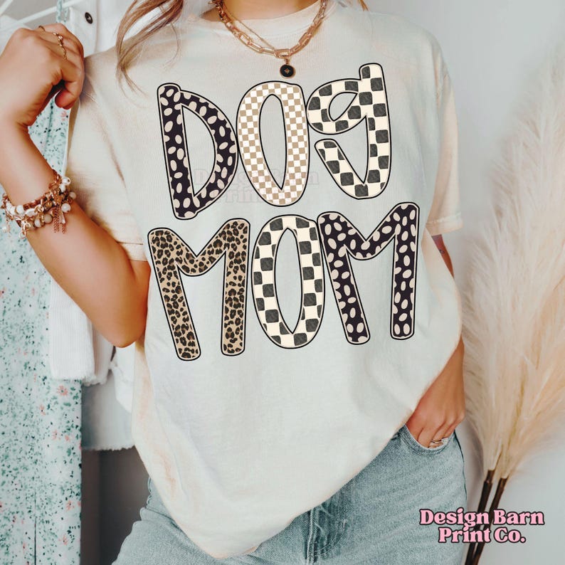 May include: Cream-colored t-shirt with the words "DOG MOM" in a bold, patterned design. The letters feature a mix of black and white polka dots, checkered patterns, and leopard print. The shirt is a casual style.