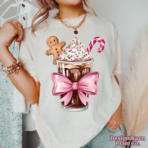 K&ouml;nnte beinhalten: Ein wei&szlig;es T-Shirt mit einem festlichen Design, das einen Lebkuchenmann, ein Schokoladengetr&auml;nk mit Schlagsahne und Streuseln sowie einen Zuckerstangen zeigt. Das Getr&auml;nk ist mit einer rosa Schleife gebunden.