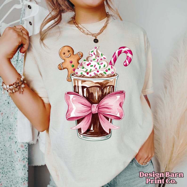 K&ouml;nnte beinhalten: Ein wei&szlig;es T-Shirt mit einem festlichen Design, das einen Lebkuchenmann, ein Schokoladengetr&auml;nk mit Schlagsahne und Streuseln sowie einen Zuckerstangen zeigt. Das Getr&auml;nk ist mit einer rosa Schleife gebunden.