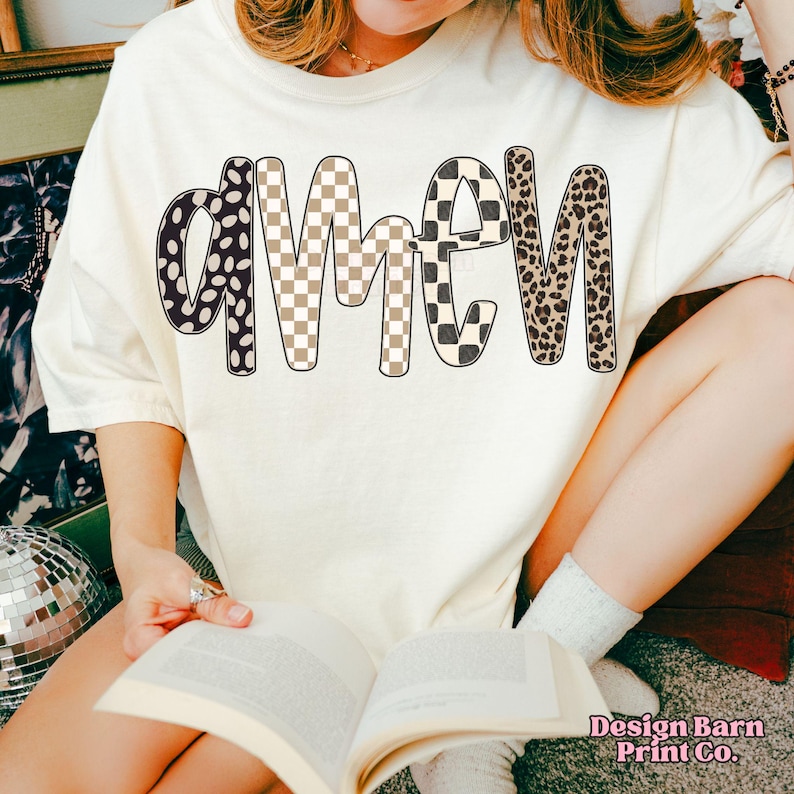 Op de afbeelding: Cr&egrave;mekleurig T-shirt met het woord "AMEN" in een gedurfd, patroonontwerp. De letters hebben stippen, dambordpatronen en luipaardprint. Het shirt maakt deel uit van een casual kledingcollectie.