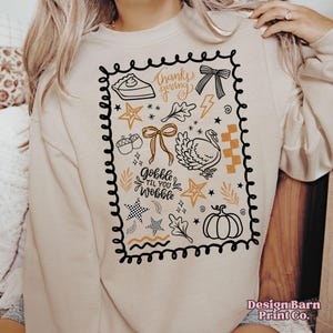 May include: Beige sweatshirt with a black and gold Thanksgiving-themed graphic. The design includes a pie, bow, turkey, stars, and the phrases "Thanksgiving" and "Gobble til you Wobble".