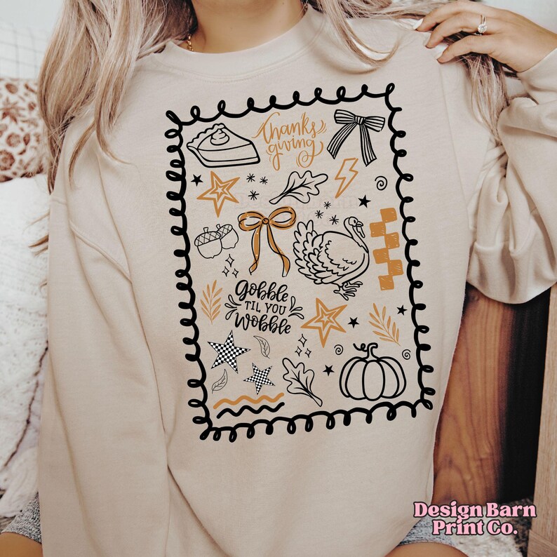 May include: Beige sweatshirt with a black and gold Thanksgiving-themed graphic. The design includes a pie, bow, turkey, stars, and the phrases "Thanksgiving" and "Gobble til you Wobble".