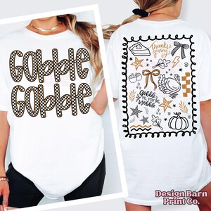 May include: White t-shirt with the words "Gobble Gobble" in a brown and black checkered pattern. The back of the shirt features Thanksgiving-themed graphics, including a turkey, pumpkin, and the phrase "Gobble til you Wobble."