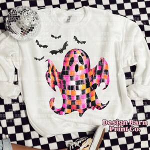 May include: White sweatshirt with a pink, orange, and black checkered ghost design. The ghost has a black outline and a black mouth. There are black bats flying around the ghost. The text "Design Barn Print Co." is printed in pink on the bottom right corner of the sweatshirt.