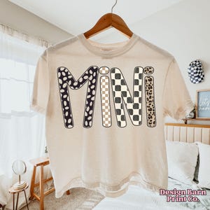 May include: Cream-colored t-shirt with the word "MINI" in a bold, playful font. The letters feature various patterns: black and white polka dots, a beige and black checkerboard, and a leopard print. The shirt is hanging on a wooden hanger.