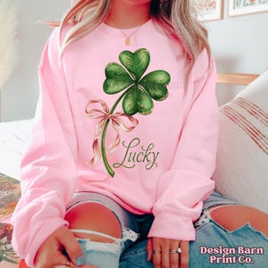Op de afbeelding: Een roze sweatshirt met een groene klaver met vier bladeren en een roze lint. De tekst "Lucky" is onder de klaver gedrukt.