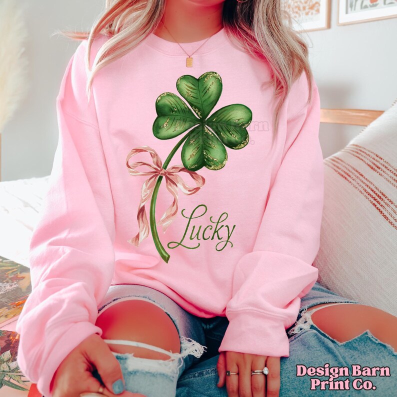 Op de afbeelding: Een roze sweatshirt met een groene klaver met vier bladeren en een roze lint. De tekst "Lucky" is onder de klaver gedrukt.