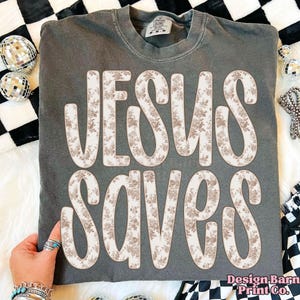 May include: A dark grey t-shirt featuring the text "JESUS SAVES" in a floral pattern. The shirt has a crew neck and short sleeves. The text is in a large, bold font with a vintage style.