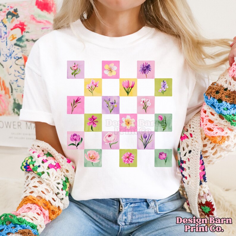 May include: White t-shirt featuring a grid of colorful floral illustrations. Each square showcases a different flower in shades of pink, purple, yellow, and green. The text "Design Barn Print Co." is visible at the bottom right.