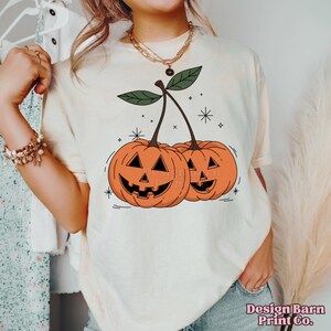 K&ouml;nnte beinhalten: Ein wei&szlig;es T-Shirt mit einem Aufdruck von zwei orangefarbenen K&uuml;rbissen mit Jack-o'-Lantern-Gesichtern, die wie Kirschen an einem Stiel aussehen.