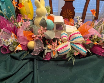 May include: A white toy truck with pink, blue, and orange ribbon bows, surrounded by pink, orange, and white mesh, and a white bunny, blue egg, and flowers. The truck is on a green tablecloth.