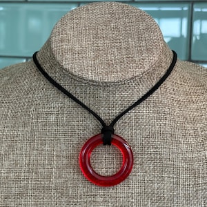 May include: A red, circular pendant necklace on a black cord. The pendant is a translucent, ring-shaped design. The necklace is displayed on a neutral-toned mannequin. The background is a light blue tile.