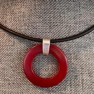 May include: A red ring pendant necklace with a silver bail, hanging from a black leather cord.