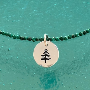 May include: A silver necklace with a round pendant featuring a small tree design. The necklace is made of green beads.