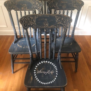 May include: Four antique chairs with carved backs painted in a distressed dark blue finish. The chairs have a simple design with a rounded back and vertical spindles. The chairs are arranged in a row with the front chair featuring a white wreath with the text "Whimsy Redo" inside.