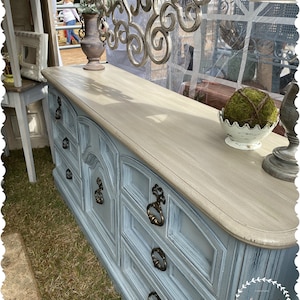 May include: A light blue dresser with six drawers and antique brass hardware. The top is a light beige color. A decorative metal scroll and a moss ball in a white bowl are on top. The dresser is on display outside.