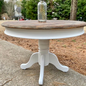May include: White round table with a wood top and a pedestal base. The table has three legs and is painted white.