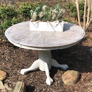 Puede incluir: Una mesa redonda blanca con un acabado desgastado y una base de pedestal con tres patas. La mesa est&aacute; decorada con una caja de plantaci&oacute;n blanca llena de vegetaci&oacute;n y flores blancas.