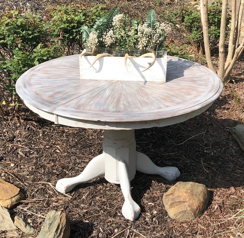 Puede incluir: Una mesa redonda blanca con un acabado desgastado y una base de pedestal con tres patas. La mesa est&aacute; decorada con una caja de plantaci&oacute;n blanca llena de vegetaci&oacute;n y flores blancas.