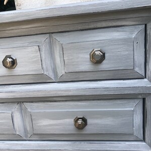 Puede incluir: Una c&oacute;moda gris con tres cajones y tiradores octogonales de plata. La c&oacute;moda tiene un acabado de pintura desgastada.