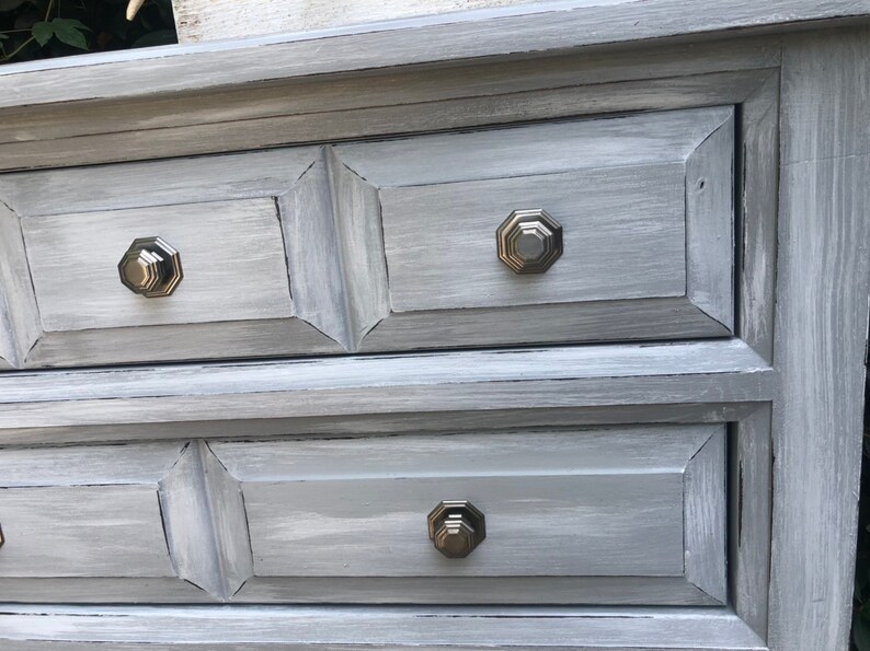Puede incluir: Una c&oacute;moda gris con tres cajones y tiradores octogonales de plata. La c&oacute;moda tiene un acabado de pintura desgastada.