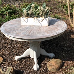 Puede incluir: Una mesa redonda blanca con un acabado desgastado y una base de pedestal con tres patas. La mesa est&aacute; decorada con una caja de plantaci&oacute;n blanca llena de flores blancas y cuernos.