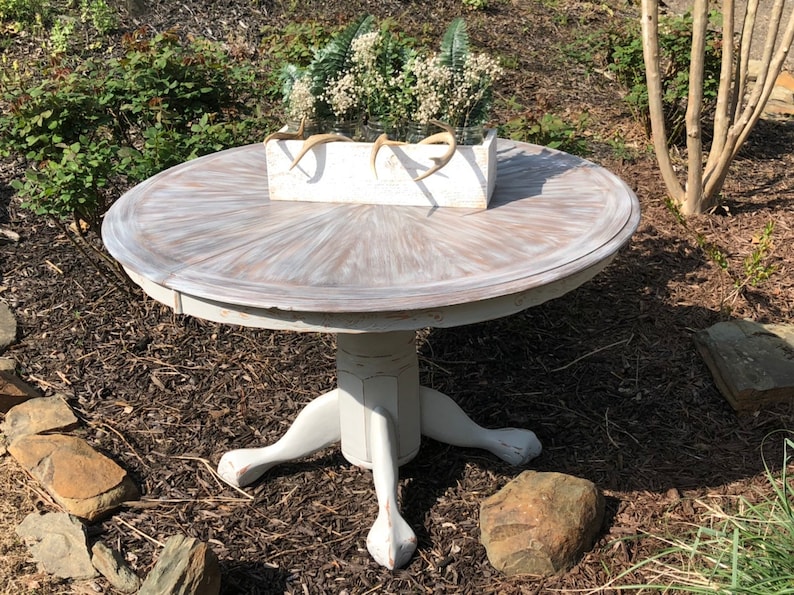 Puede incluir: Una mesa redonda blanca con un acabado desgastado y una base de pedestal con tres patas. La mesa est&aacute; decorada con una caja de plantaci&oacute;n blanca llena de flores blancas y cuernos.