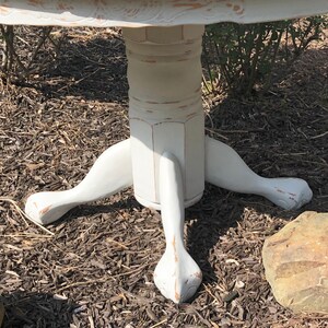 Puede incluir: Mesa redonda pintada de blanco con una base de pedestal y tres patas. Las patas est&aacute;n talladas con un dise&ntilde;o de pata de garra. La mesa est&aacute; envejecida y tiene un aspecto r&uacute;stico.