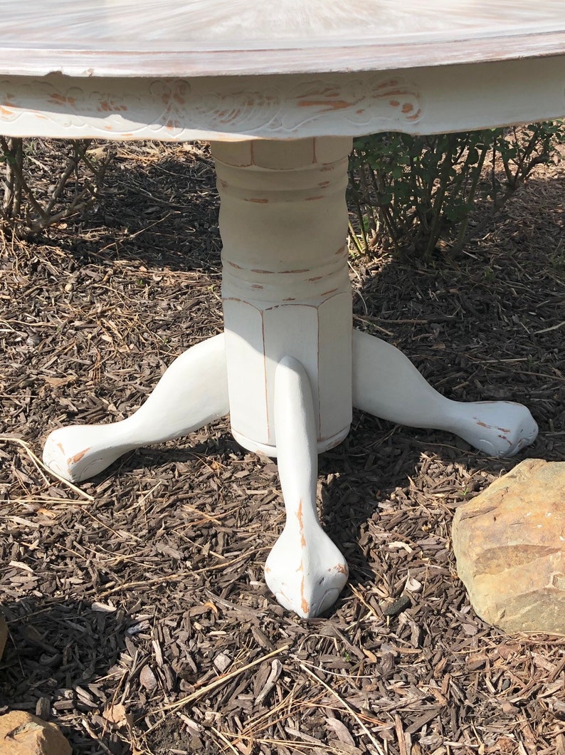 Puede incluir: Mesa redonda pintada de blanco con una base de pedestal y tres patas. Las patas est&aacute;n talladas con un dise&ntilde;o de pata de garra. La mesa est&aacute; envejecida y tiene un aspecto r&uacute;stico.
