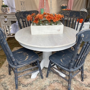 May include: A white round table with a distressed white finish and four black chairs with distressed black paint. The table has a pedestal base and the chairs have a spindle back. A wooden box with orange flowers sits on the table.