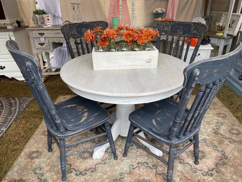 May include: A white round table with a distressed white finish and four black chairs with distressed black paint. The table has a pedestal base and the chairs have a spindle back. A wooden box with orange flowers sits on the table.