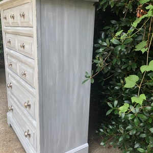 Puede incluir: Una c&oacute;moda de madera pintada de gris con seis cajones y tiradores de cajones plateados. La c&oacute;moda tiene un acabado desgastado y est&aacute; sobre una superficie de hormig&oacute;n.
