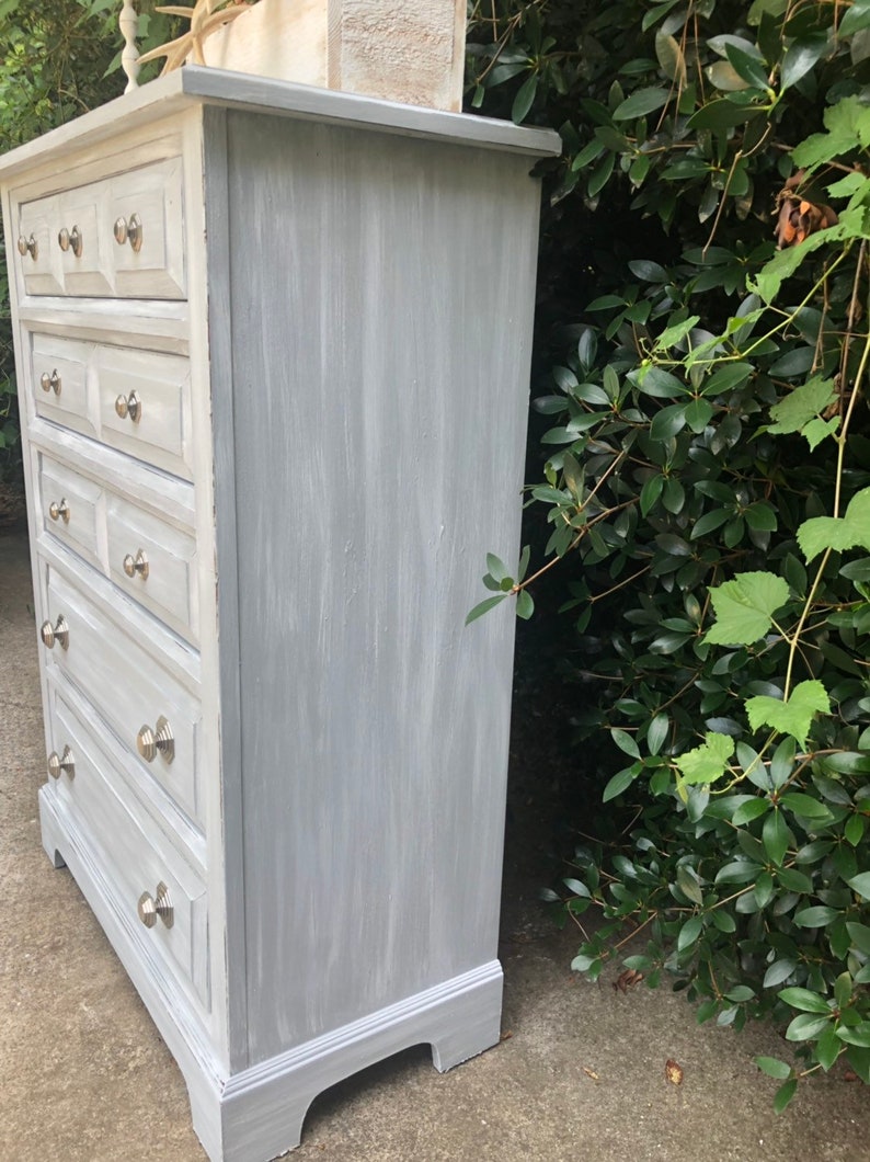 Puede incluir: Una c&oacute;moda de madera pintada de gris con seis cajones y tiradores de cajones plateados. La c&oacute;moda tiene un acabado desgastado y est&aacute; sobre una superficie de hormig&oacute;n.