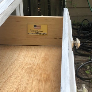 Puede incluir: Primer plano de un caj&oacute;n con un interior de madera y un exterior pintado de blanco. El caj&oacute;n tiene una etiqueta dorada con el texto "Vaughan-Bassett" y una bandera estadounidense.