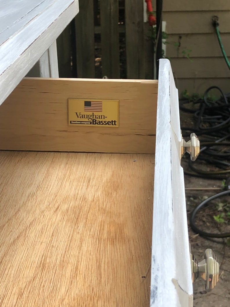 Puede incluir: Primer plano de un caj&oacute;n con un interior de madera y un exterior pintado de blanco. El caj&oacute;n tiene una etiqueta dorada con el texto "Vaughan-Bassett" y una bandera estadounidense.