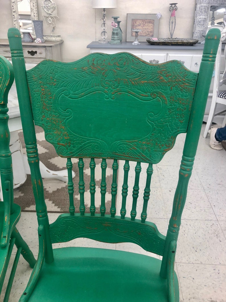 Pu&ograve; includere: Una sedia in legno verde con schienale intagliato e finitura usurata. La sedia ha uno schienale a fuso e una parte superiore curva.